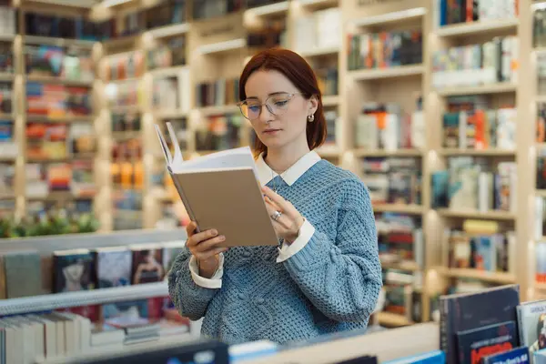 Kütüphane rafları arasında kitap okumaya odaklanmış bir kadın..