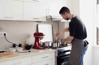 Adam temiz bir mutfakta yemek pişiriyor, soba kullanıyor ve kırmızı bir karıştırıcı kullanıyor..