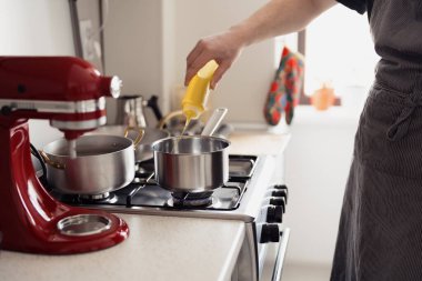 Mutfakta yemek pişiren, gaz ocağındaki tencereye yağ ekleyen ve yanında kırmızı bir mikser olan biri..