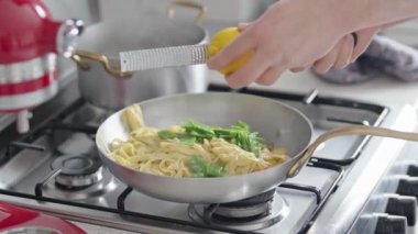 Erişteli ve yeşil sebzeli bir tencere üzerinde limon ızgarası yapan ellerin yakın çekimi modern bir mutfakta ev yapımı sağlıklı yemek hazırlığını vurguluyor..