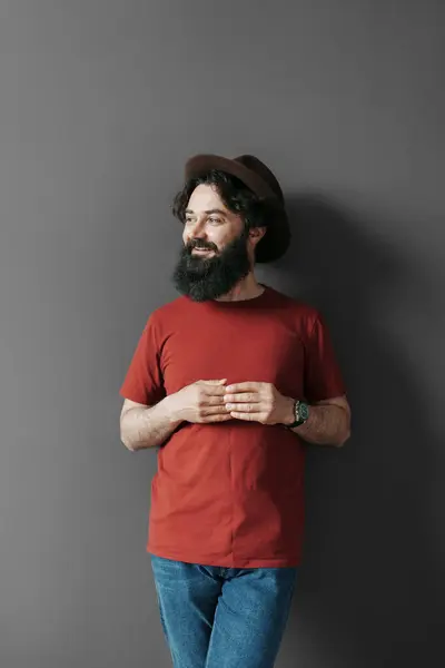 温かい笑顔のスタイリッシュなひげの男のカジュアルな肖像画 赤いTシャツを着て グレーの背景にグレーの帽子 ジーンズを着て 自信とレイドバックの魅力を除いて ロイヤリティフリーのストック写真
