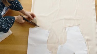 Terzilerin yakın plan fotoğrafları. Elleri titizlikle ahşap bir yüzey üzerinde bej kumaş kesiyor. Kağıt desenli..