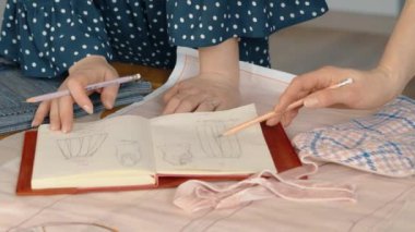 Moda tasarımcılarının elleri, kumaşla çevrili bir deftere kıyafet tasarımları çizerken..