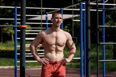 Fiziki olarak fit bir adam açık hava spor salonunda kaslı, kaslı vücudunu parlak bir günde spor aletlerine karşı sergiliyor..