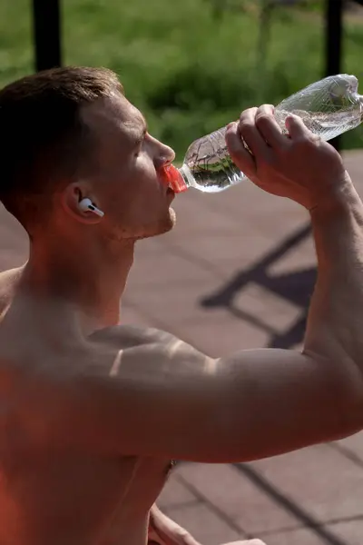 A fit, athletic man takes a refreshing drink from a clear water bottle, exemplifying health and fitness during an outdoor workout session.