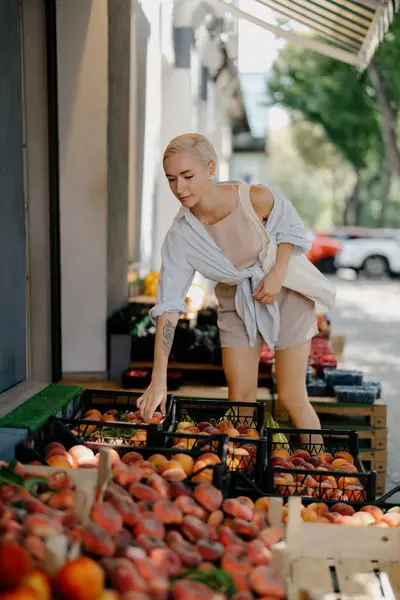 スタイリッシュな若い女性は 日当たりの良い農家市場でクレートから新鮮な桃を慎重に選択します ロイヤリティフリーのストック写真