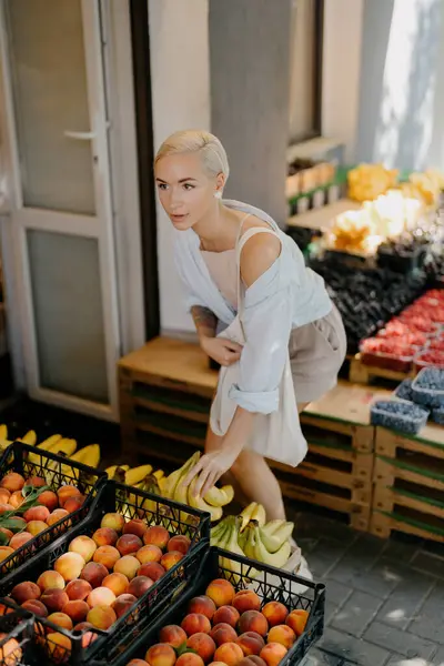 短いブロンドの髪を持つスタイリッシュな若い女性は晴れた日に活気に満ちたマーケットストールから新鮮なフルーツを選択します ストック画像