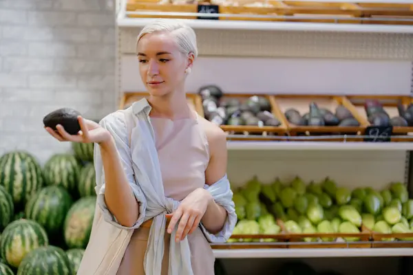 若い女性店員が新鮮な果物に囲まれた食料品店の農産物セクションで思慮深い表情でアボカドを調べる ロイヤリティフリーのストック画像