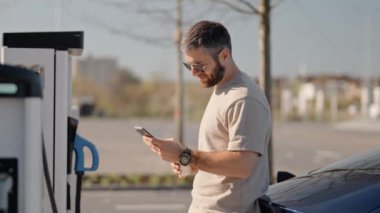 Orta yaşlı bir adam, güneşli bir günde modern bir açık hava şarj istasyonunda elektrikli arabası şarj olurken akıllı telefonunu kontrol eder..