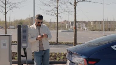 Sıradan giyinmiş bir adam, güneşli bir günde elektrikli arabasının şarj olmasını sabırla beklerken telefonunu kontrol eder..