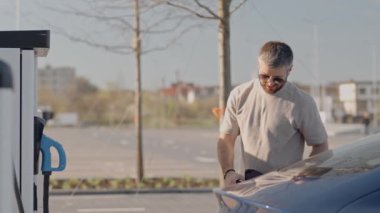 Genç bir erkek elektrikli aracını şarj istasyonunda fişini çekerken yakalanır, aynı anda akıllı telefonunu kullanır, muhtemelen programını yönetir ya da bildirimlerini kontrol eder..