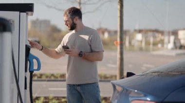 Yetişkin erkek, güneşli bir günde elektrikli araç EV şarj istasyonunu izlemek ya da işletmek için akıllı telefon kullanıyor, modern, çevre dostu teknoloji sergiliyor..