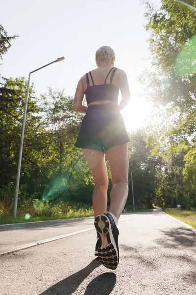 Back View Woman Jogging Sunlit Road Surrounded Greenery Showcasing Active 图库图片