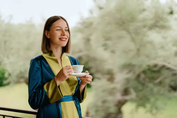 穏やかでコンテンツの若い女性は 緑豊かな緑に囲まれた笑顔でコーヒーを飲むバルコニーの上に立っています ストックフォト