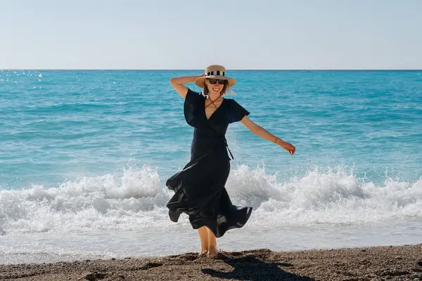 黒いドレスと太陽の喜びの女性は 晴れた晴れた日に海の波に遊んで飛び散り レジャーと幸せを体現します ストック画像