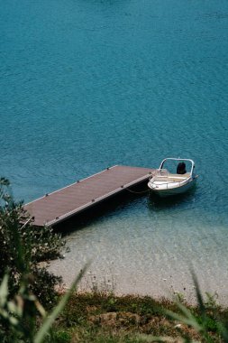 Yüzen ahşap bir rıhtıma bağlı, temiz su ve kıyı yeşillikleriyle çevrili küçük bir teknenin yer aldığı sakin bir göl..