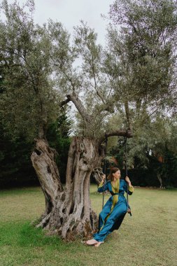 Mavi elbiseli, yaşlı bir zeytin ağacına bağlı kırsal bir salıncakta sallanan genç bir kadının huzurlu görüntüsü doğada sakinlik ve rahatlama hissi uyandırıyor..