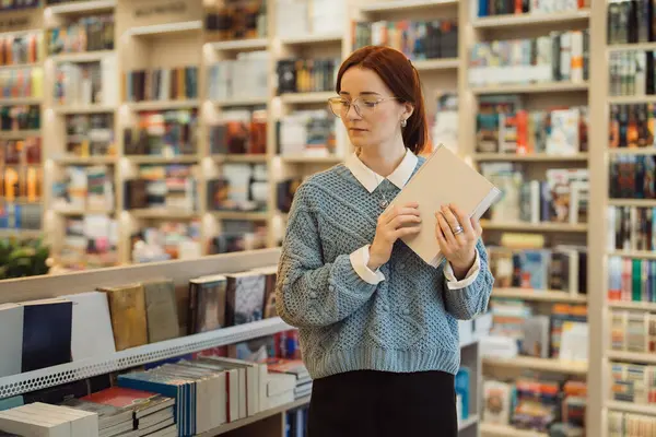 아늑한 스웨터를 여성은 문학이 가득한 선반에 둘러싸인 갖춰진 도서관에서 있습니다 스톡 사진