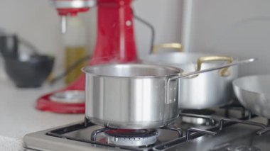 Yakından çekilen bir görüntü, gaz sobasındaki yemek tenceresinden çıkan buharı yakalıyor, temiz, çağdaş bir mutfağın arka planına kurulmuş, çeşitli mutfak aletleri ve canlı kırmızı bir mikserle..