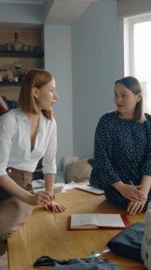 İki kadın moda tasarımcısı, masanın üzerinde malzeme ve çizimler olan parlak bir atölyede iş birliği yapıyor ve tasarımları tartışıyorlar..