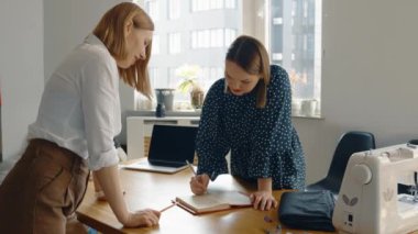 İki kadının, iyi aydınlatılmış bir tasarım stüdyosunda, dikiş ekipmanları ve dizüstü bilgisayarla çevrili bir proje üzerinde tartışırken çekilmiş yaratıcı bir işbirliği anı..