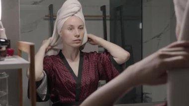 Kırmızı bornozlu ve kafasında havlu olan bir kadın banyo aynasında kendine bakıyor..