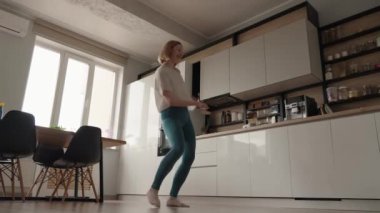 Genç bir kadın modern mutfağında mutlu bir şekilde dans ediyor, eğlenceli ve kaygısız bir anın tadını çıkarıyor. Parlak ve minimalist iç mimari.