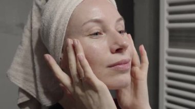 Kafasında havlu olan bir kadın duştan sonra yüzüne krem sürüyor, kendine bakmasını ve güzellik alışkanlıklarını teşvik ediyor..