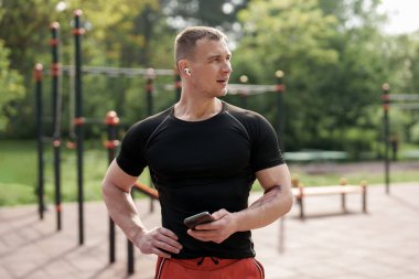 Formda, atletik bir adam spor yaptıktan sonra, akıllı telefonunu tutuyor ve kablosuz kulaklık takıyor. Açık hava fitness parkında duruyor, etkin bir yaşam tarzı sergiliyor..