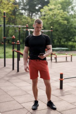Atletik bir adam açık hava spor salonunda duruyor, akıllı telefonuna odaklanmış. Üzerinde siyah tişört ve kırmızı şort vardı. Huzurlu park ortamının ortasında spor yapmaya hazır görünüyordu..