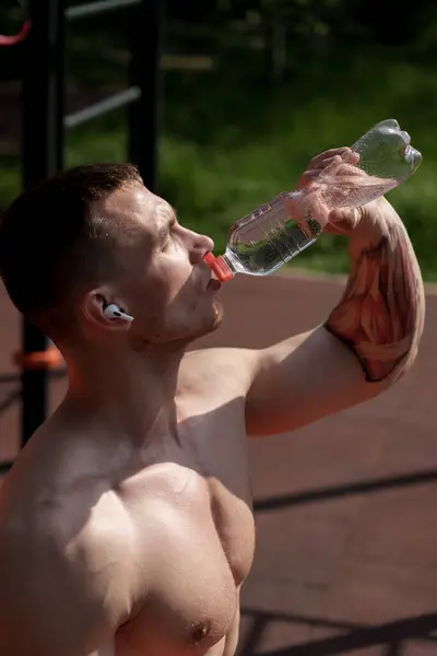 stock image An athletic man pours water over his face to cool down after an intense workout on a sunny day in the park. The focus is on fitness and refreshment.