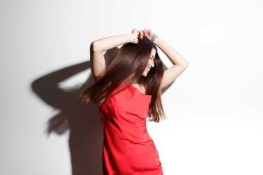 Parlak kırmızı elbiseli, elleri saçlarında, beyaz bir arka plana yaslanmış neşeli genç bir kadının canlı görüntüsü mutluluk ve güven saçıyor..