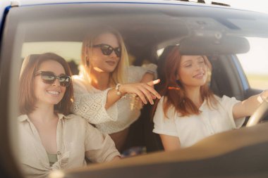 Üç neşeli kadın, araba gezintisi sırasında gülüşlerini aydınlatan sıcak güneş ışığıyla eğlenceli bir anı paylaşıyorlar..