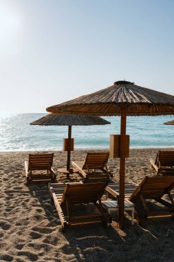 Güneşli sahil manzarası, ahşap güneş şezlongları ve kumlu sahilde saman şemsiyeleri denize bakıyor..