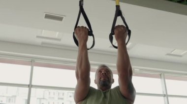 Modern bir spor salonunda askılarla çalışan olgun bir adam. Güç, zindelik ve adanmışlık gösteriyor. Parlak, iyi ışıklandırılmış, geniş pencereli..