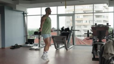 Geniş pencereli ve şehir manzaralı modern bir spor salonunda ip atlayan bir adam. Odaklanmış ve fiziksel kondisyonunu koruyor..