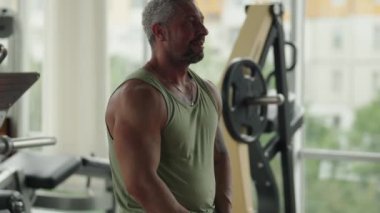 Spor salonundaki kaslı bir adam, güç egzersizleri yapmaya hazır. Yeşil bir atlet giyiyor, formda fiziğini ve formda kalma kararlılığını gösteriyor..