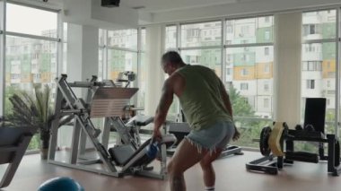 Modern bir spor salonunda bir adam salıncak zili ile salıncak egzersizi yapıyor. Spor salonunda doğal ışık sağlayan geniş pencereli çeşitli ekipmanlar yer alıyor..