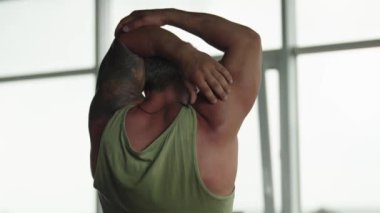 Yeşil kolsuz tişörtlü kaslı bir adam, parlak bir spor salonunda yoğun bir antrenmandan sonra kollarını esnetiyor..