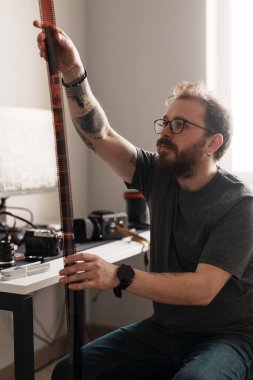 Odaklanmış sanatçı, fotoğraf ekipmanlarıyla çevrili parlak ve modern bir çalışma alanında film şeritlerini dikkatle tutuyor ve inceliyor..