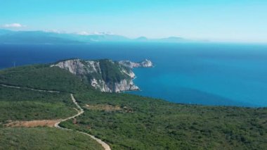 Canlı bir kıyı şeridi manzarası, yeşil tepeler, kayalık uçurumlar, ve sakin mavi bir denizin arka planına uzanan dolambaçlı bir yol..