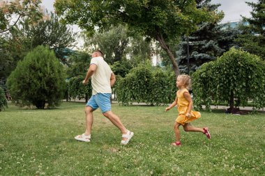 Baba ve kızı yeşil bir parkta koşuyor, güneşli bir günde aile ve açık havada eğleniyorlar..