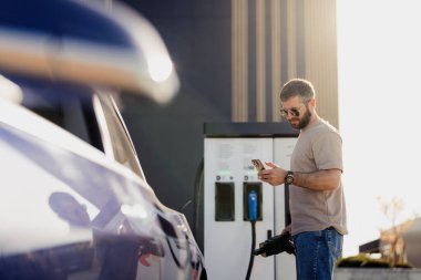 Yetişkin erkek, güneşli bir günde telefonunu kontrol ederken elektrikli aracı şarj istasyonuna bağlar..