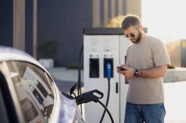 Orta yaşlı bir adam, güneşli bir günde elektrikli arabası şarj olurken akıllı telefonunu kontrol eder..