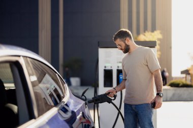 Şarj cihazını şarj istasyonuna takan odaklanmış bir adamın yan görüntüsü. Arka planda modern şehir mimarisi var..