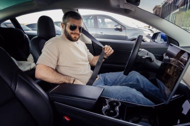 Güneş gözlüklü şık bir adam emniyet ve rahatlığı vurgulayarak çağdaş bir elektrikli aracın içinde gülümserken emniyet kemerini bağlar..