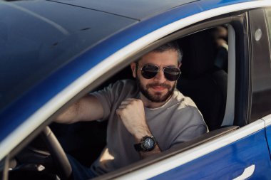 Güneş gözlüklü neşeli sakallı bir adam güneşli bir günün keyfini çıkarırken mavi elektrikli bir arabanın modern ve gösterişli kamarasında oturuyor..