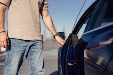Modern mavi elektrikli bir arabanın şoför tarafındaki kapısını açan bir adamın yakın plan görüntüsü, çevre dostu bir ulaşım sergiliyor..