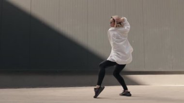 Beyaz gömlekli ve siyah pantolonlu genç bir kadın güneşte dans ediyor, hareketleri akıcı ve anlamlı. Minimalist arka plan ve doğal ışıklandırma şehirli şıklığı yaratıyor..