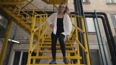Zarif genç bir kadın sarı endüstriyel bir merdivende güven içinde dans ediyor. Hareketleri enerjik ve anlamlı, dans tutkusunu ve neşesini gösteriyor..